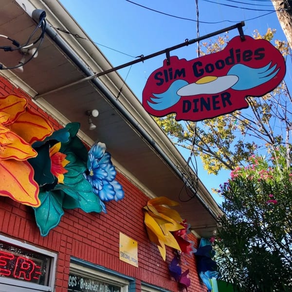 Slim Goodies Diner sign, featuring a winged fried egg, hangs above a red brick building with colorful flower decorations.