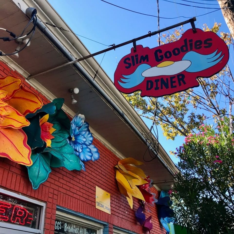 Slim Goodies Diner sign, featuring a winged fried egg, hangs above a red brick building with colorful flower decorations.