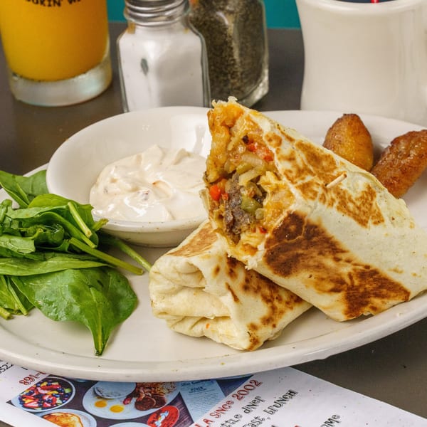 A breakfast burrito, cut in half, with a side of spinach, dipping sauce, and plantains.
